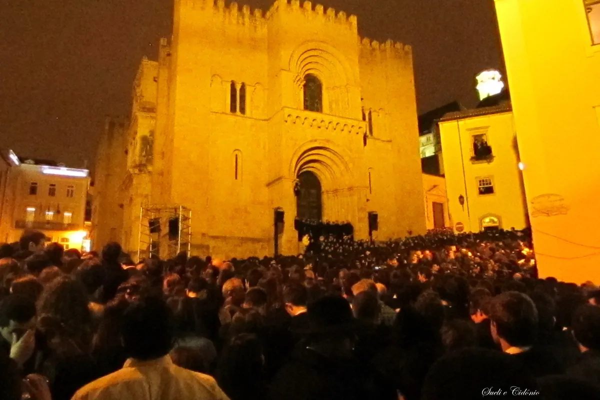 A large crowd stands in the dark in front of the lit stone front of Coimbra's Old Cathedral during a night serenade