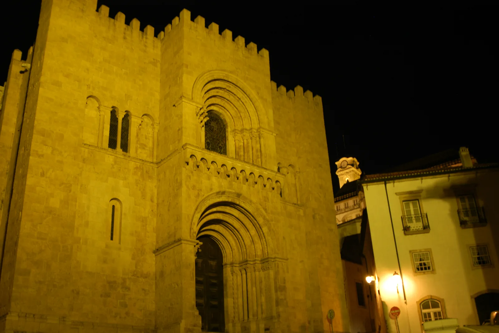 The crenellated Romanesque front of Coimbra's Old Cathedral, lit gold against a night sky, with lit houses at the edge of the square