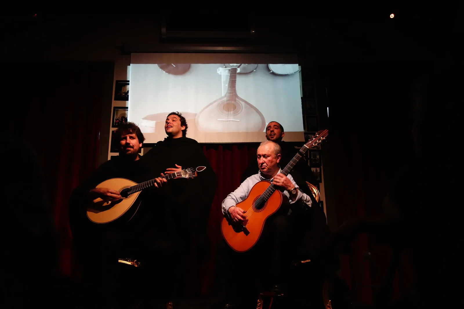 A singer with eyes half closed stands behind a Coimbra guitarist and a classical guitarist, all in black capes, in a dim room with red curtains