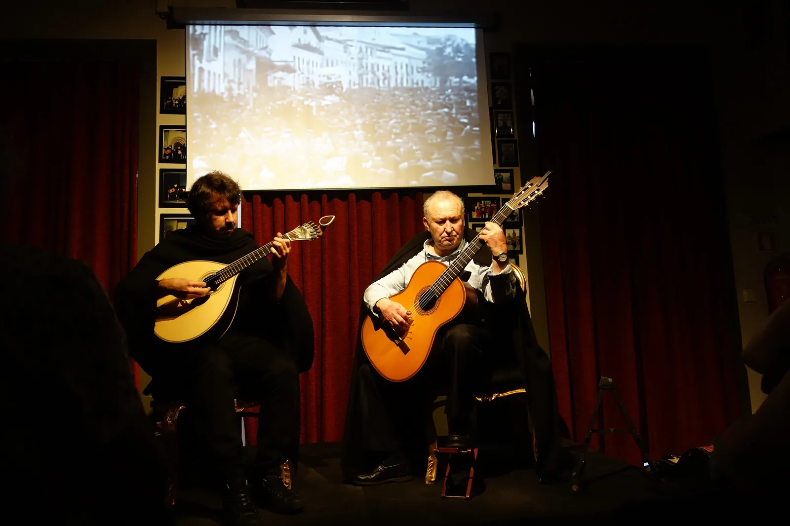 Two musicians in black capes sit on a small stage, one with a Coimbra guitar and one with a classical guitar, in front of red curtains and a projection screen